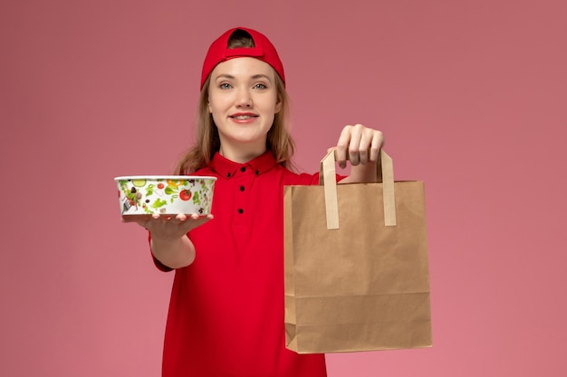 Kostenloses Foto vorderer blick junger weiblicher kurier in roter uniform, die lieferung nahrungsmittelpaket und schüssel auf der hellrosa wand hält