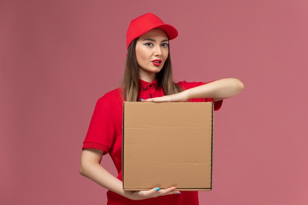 Vorderer Blick junger weiblicher Kurier in roter Uniform, die Nahrungsmittelbox auf rosa Hintergrundarbeiterservice-Lieferuniform-Firmenjob hält