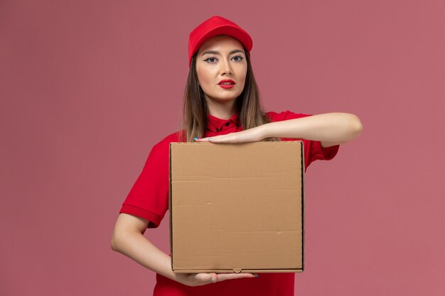 Vorderer Blick junger weiblicher Kurier in roter Uniform, die Nahrungsmittelbox auf rosa Hintergrundarbeiterservice-Lieferuniform-Firmenjob hält