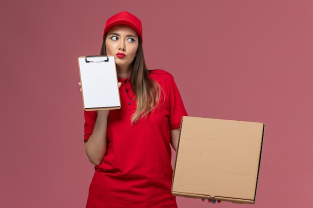 Vorderer Blick junger weiblicher Kurier in roter Uniform, die Nahrungsmittelbox mit Notizblock hält und auf rosa Hintergrundlieferdienstuniformfirmenjobarbeit denkt