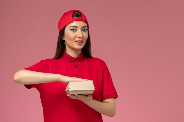 Vorderer Blick junger weiblicher Kurier in roter Uniform und Umhang mit wenig Liefernahrungsmittelpaket auf ihren Händen an der rosa Wand