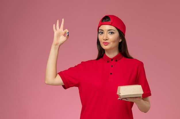 Vorderer Blick junger weiblicher Kurier in roter Uniform und Umhang mit wenig Liefernahrungsmittelpaket auf ihren Händen an der rosa Wand