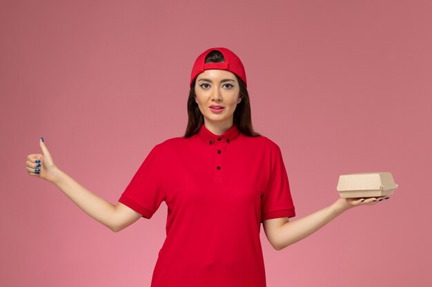 Vorderer Blick junger weiblicher Kurier in roter Uniform und Umhang mit wenig Liefernahrungsmittelpaket auf ihren Händen an der rosa Wand