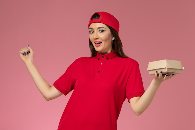 Vorderer Blick junger weiblicher Kurier in roter Uniform und Umhang mit wenig Liefernahrungsmittelpaket auf ihren Händen an der rosa Wand