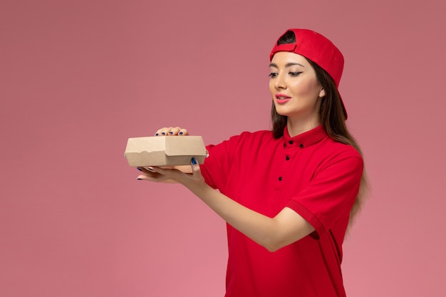 Vorderer Blick junger weiblicher Kurier in roter Uniform und Umhang mit wenig Liefernahrungsmittelpaket auf ihren Händen auf hellrosa Wand