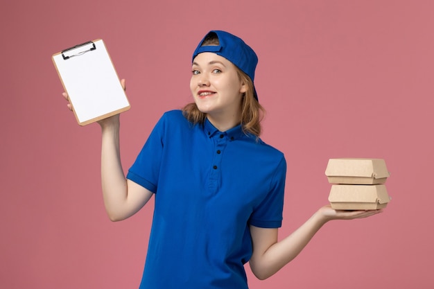 Vorderer Blick weiblicher Kurier in blauem Uniformumhang, der kleine Liefernahrungsmittelpakete und Notizblock auf rosa Hintergrundlieferdienstmitarbeiter-Arbeiterjob hält