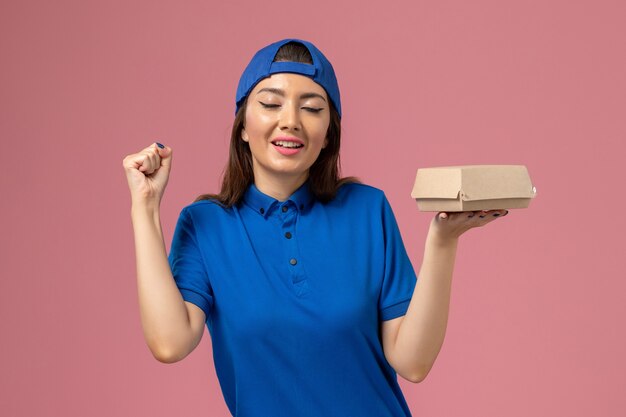 Vorderer Blick weiblicher Kurier in blauem Uniformumhang, der kleines Lieferpaket hält und sich auf rosa Wand freut, Mitarbeiterdienst-Lieferarbeitsjob