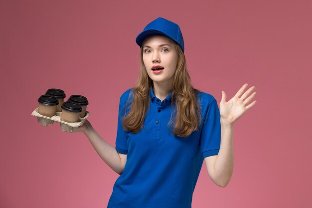 Vorderer Blick weiblicher Kurier in blauer Uniform, die braune Lieferkaffeetassen mit überraschtem Ausdruck auf dem rosa Schreibtischdienstuniformfirmenarbeiter hält
