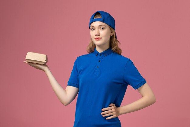 Vorderer Blick weiblicher Kurier in der blauen Uniform und im Umhang, die kleines Liefernahrungsmittelpaket auf rosa Hintergrundjoblieferuniformuniversitätsarbeiterfirma halten