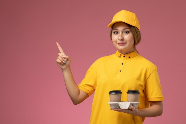 Vorderer Blick weiblicher Kurier in gelbem einheitlichem gelbem Umhang, der kunststoffbraune Kaffeetassen auf der weiblichen Farbe der rosa Hintergrunduniformlieferung hält