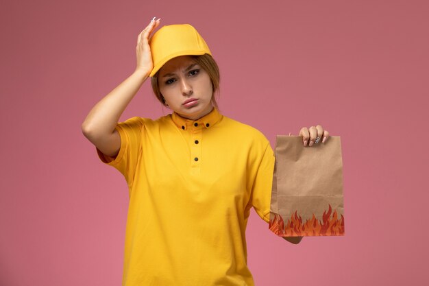 Vorderer Blick weiblicher Kurier in gelbem einheitlichem gelbem Umhang, der Lebensmittelpaket mit starkem Kopfschmerz rosa Hintergrunduniformlieferarbeitsfarbjob hält