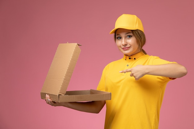 Vorderer blick weiblicher kurier in gelbem einheitlichem gelbem umhang, der leere lebensmittelverpackung auf dem rosa hintergrunduniformlieferungsarbeitsjob hält