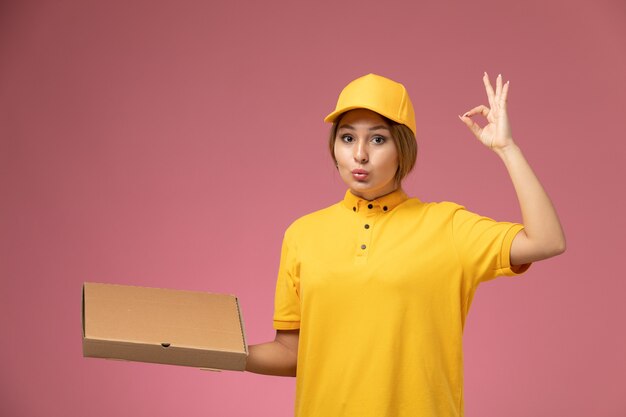Vorderer Blick weiblicher Kurier in gelbem einheitlichem gelbem Umhang, der Nahrungsmittelbox-Paket auf rosa Schreibtischuniformlieferung weiblicher Arbeitsfarbe hält