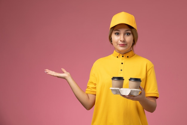 Vorderer Blick weiblicher Kurier in gelbem einheitlichem gelbem Umhang, der Plastikkaffeetassen auf dem rosa Hintergrunduniformlieferungsarbeitsjob hält