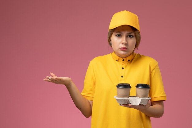 Vorderer Blick weiblicher Kurier in gelbem einheitlichem gelbem Umhang, der Plastikkaffeetassen auf der rosa Hintergrunduniformlieferarbeitsjobfarbe hält