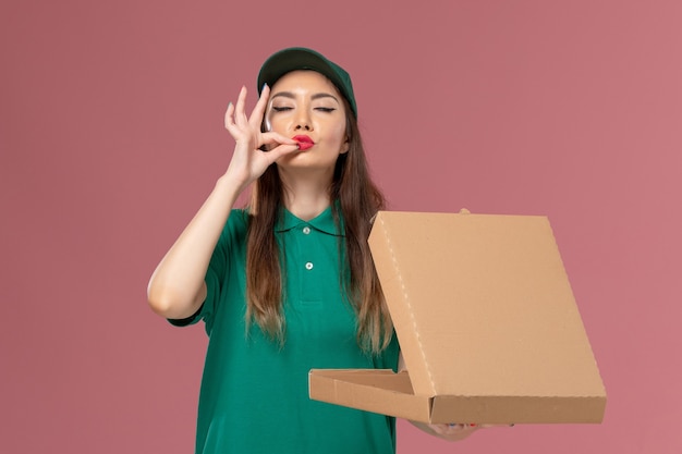 Vorderer Blick weiblicher Kurier in grüner Uniform, die Lebensmittellieferbox auf rosa Wanddienstuniform-Liefermädchenfirma hält und öffnet