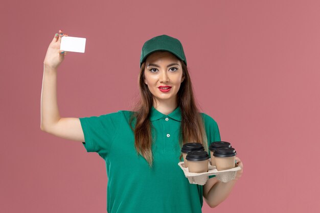 Vorderer Blick weiblicher Kurier in grüner Uniform und Umhang, der Karte und Lieferkaffeetassen auf rosa Wandserviceuniform-Lieferauftragsarbeit hält