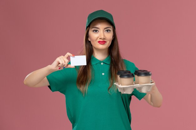 Vorderer Blick weiblicher Kurier in grüner Uniform und Umhang, der Karte und Lieferkaffeetassen auf rosa Wandserviceuniformlieferauftrag hält