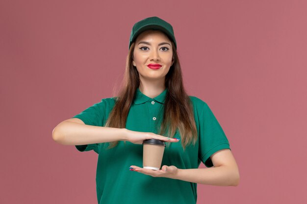 Vorderer Blick weiblicher Kurier in grüner Uniform und Umhang, der Lieferung Kaffeetasse auf rosa Wand Firmenservice Jobuniform Lieferarbeiter weibliche Arbeit hält