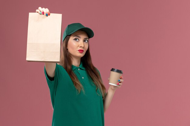Vorderer Blick weiblicher Kurier in grüner Uniform und Umhang, der Lieferung Kaffeetasse und Lebensmittelpaket auf rosa Wand Firmenservice Jobuniform Lieferarbeiter weibliche Arbeit hält