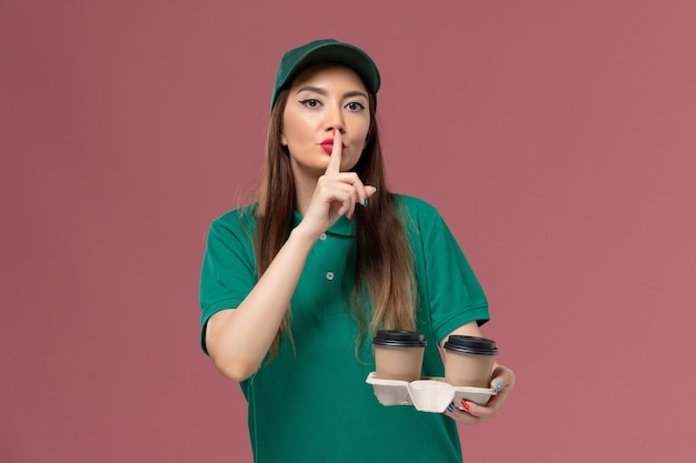 Vorderer Blick weiblicher Kurier in grüner Uniform und Umhang, der Lieferung Kaffeetassen auf rosa Wand Firmenservice Uniform Lieferung weibliches Arbeitsmädchen hält