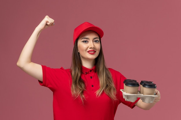 Vorderer Blick weiblicher Kurier in roter Uniform, die Lieferkaffeetassen hält, die auf rosa Hintergrunddienstlieferungsuniform-Jobarbeiter flüstern