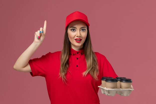 Vorderer Blick weiblicher Kurier in roter Uniform, die Lieferkaffeetassen hält, die ihren Finger auf rosa Hintergrundarbeiterjobdienstlieferuniform heben