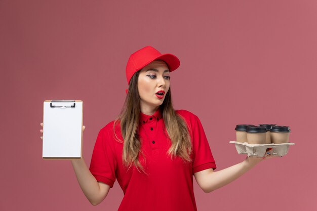 Vorderer Blick weiblicher Kurier in roter Uniform, die Lieferkaffeetassen und Notizblock auf rosa Bodenservice-Lieferfirmenjobuniform hält