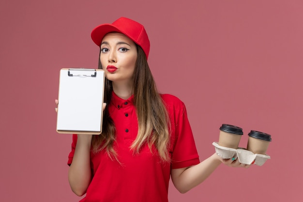 Vorderer Blick weiblicher Kurier in roter Uniform, die Lieferkaffeetassen und Notizblock auf rosa Hintergrunddienstlieferungsuniform hält