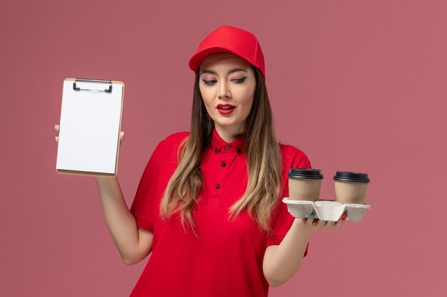 Vorderer Blick weiblicher Kurier in roter Uniform, die Lieferkaffeetassen und Notizblock auf rosa Schreibtischdienstlieferungsjobarbeiteruniform hält