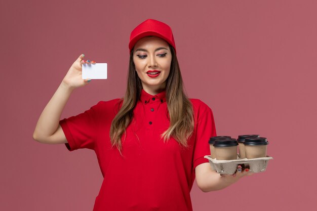 Vorderer Blick weiblicher Kurier in roter Uniform, die Lieferkaffeetassen und Plastikkarte auf rosa Hintergrunddienstlieferungsjobuniform hält