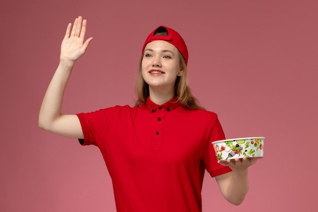 Vorderer Blick weiblicher Kurier in roter Uniform und Umhang, der Lieferschüssel auf hellrosa Wand hält, Arbeiterjobmädchenserviceuniformlieferung