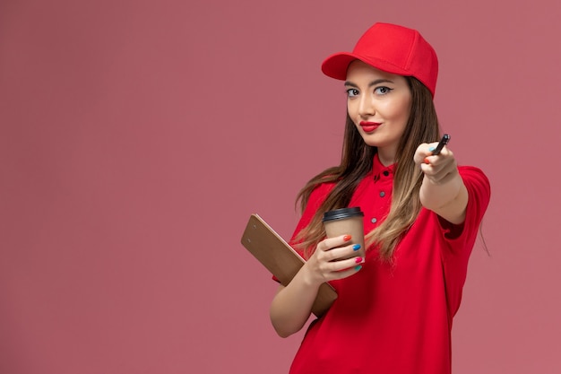 Vorderer Blick weiblicher Kurier in roter Uniform und Umhang, der Lieferung Kaffeetasse Notizblock und Stift auf rosa Hintergrund Arbeiter Service Lieferung Uniform Job hält
