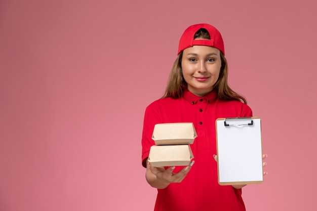 Vorderer Blick weiblicher Kurier in roter Uniform und Umhang, der Notizblock und kleine Liefernahrungsmittelpakete auf rosa Hintergrundjobuniform-Lieferservice hält