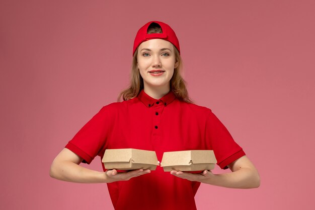 Vorderer Blick weiblicher Kurier in roter Uniform und Umhang, die kleine Liefernahrungsmittelpakete mit Lächeln auf rosa Wand halten, Arbeitslieferdienstunternehmenuniformjob