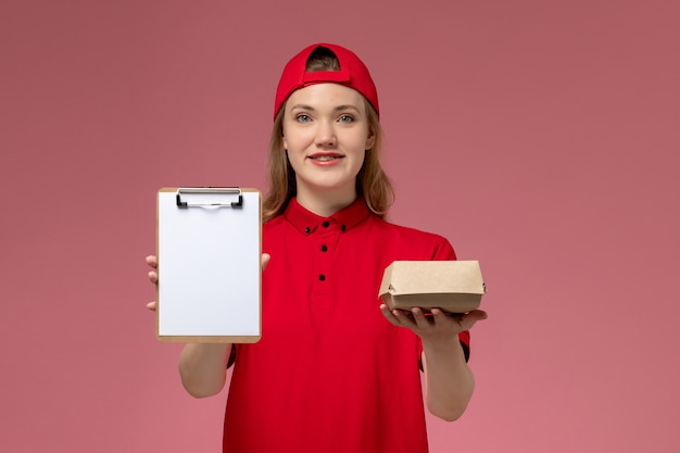 Vorderer Blick weiblicher Kurier in roter Uniform und Umhang, die kleines Liefernahrungsmittelpaket mit Notizblock auf der rosa Wand, Jobuniform des Lieferserviceunternehmens halten