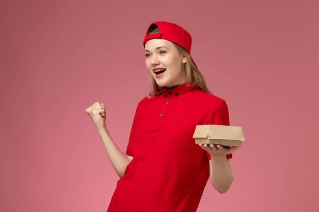 Vorderer Blick weiblicher Kurier in roter Uniform und Umhangjubel, der kleines Liefernahrungsmittelpaket auf rosa Wand, Uniformdienst des Lieferserviceunternehmens hält