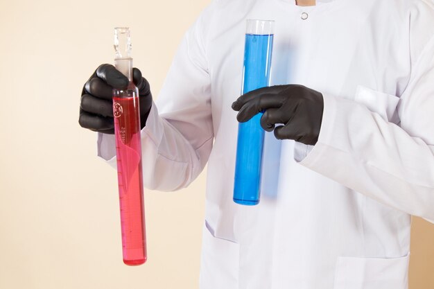 Vorderer Nahansicht junger männlicher Chemiker im weißen Spezialanzug, der kleine Flaschen mit farbigen Lösungen auf Cremewandchemie-Wissenschaftslaborexperiment hält