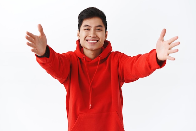 Waistup-Porträt eines hübschen, attraktiven asiatischen Mannes in rotem Hoodie, der die Hände in Richtung Kamera streckt, möchte einen herzlichen Willkommensfreund mit Kuscheln geben, der auf breitem weißem Hintergrund lächelt