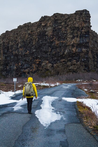 Wanderer in einer gelben Jacke, die durch die Straße geht, umgeben von Felsen und einem Feld in Island