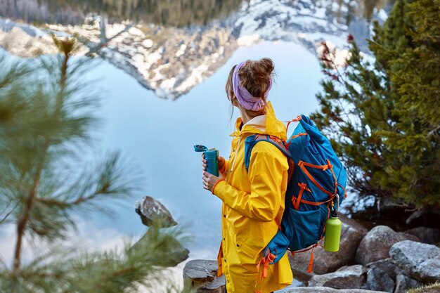 Wanderfrau hält in der Nähe des Sees in den Bergen an, trägt einen Rücken, hält eine Thermoskanne mit Heißgetränken und erkundet etwas Neues