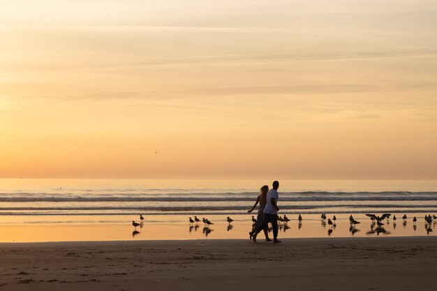 Warmes Paar, das bei Sonnenuntergang am Strand spazieren geht. Mann und Frau in Freizeitkleidung, die in der Abenddämmerung am Wasser entlang laufen. Liebe, Familie, Naturkonzept