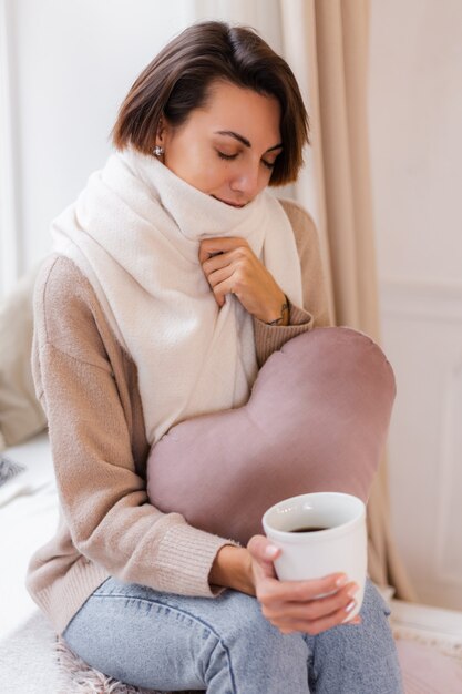 Warmes Porträt der Frau, die auf Fensterbank mit Tasse heißem Teekaffee sitzt, der Pullover und weißen Schal trägt