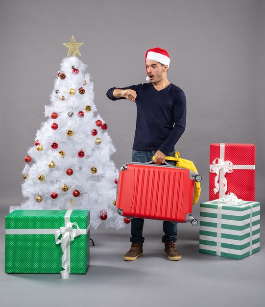 wartender junger Mann mit Weihnachtsmütze nahe weißem Weihnachtsbaum auf grau