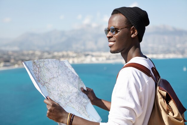 Was für eine wunderschöne Landschaft! Glücklicher aufgeregter afroamerikanischer Rucksacktourist, der Papierkarte verwendet, während er auf Aussichtspunkt hoch über blauem Meer steht und Umgebung während seiner Reise studiert. Reisen und Abenteuer