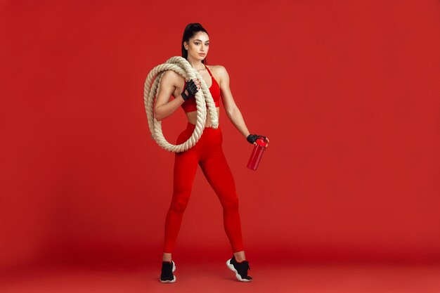 Wasser trinken. Schöne junge Sportlerin, die in einfarbigem rotem Porträt übt. Brünettes Modell mit sportlicher Passform mit Seilen. Bodybuilding, gesunder Lebensstil, Schönheits- und Aktionskonzept.