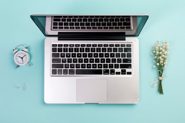 Wecker, Büroklammer, Maiglöckchenblumenstrauß mit einem offenen Laptop auf blauem Geschäftsschreibtisch