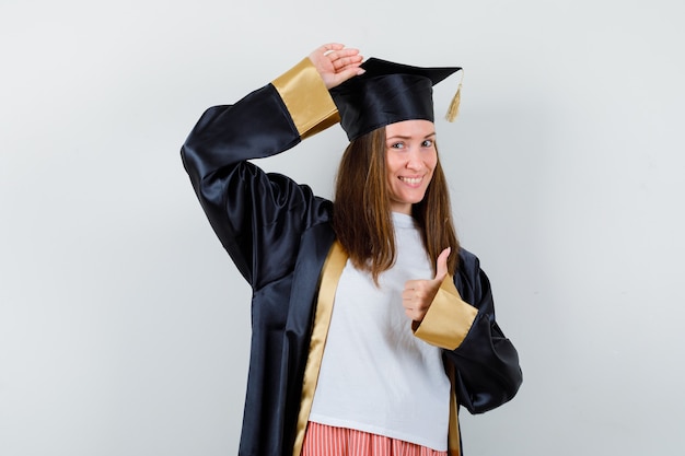 Weibliche Absolventin zeigt Daumen hoch, hält Hand auf Kopf in Uniform, Freizeitkleidung und sieht glücklich aus, Vorderansicht.