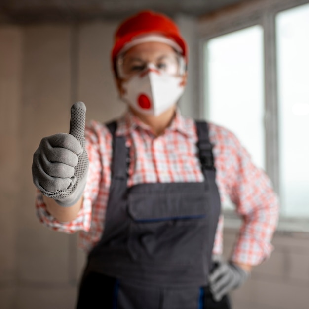 Kostenloses Foto weibliche bauarbeiterin mit helm und gesichtsmaske zeigt daumen hoch