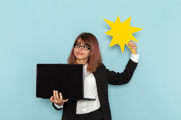 Weibliche Büroangestellte der Vorderansicht im strengen Anzug unter Verwendung des gelben Zeichens des Laptops, der auf hellblauer Oberfläche hält